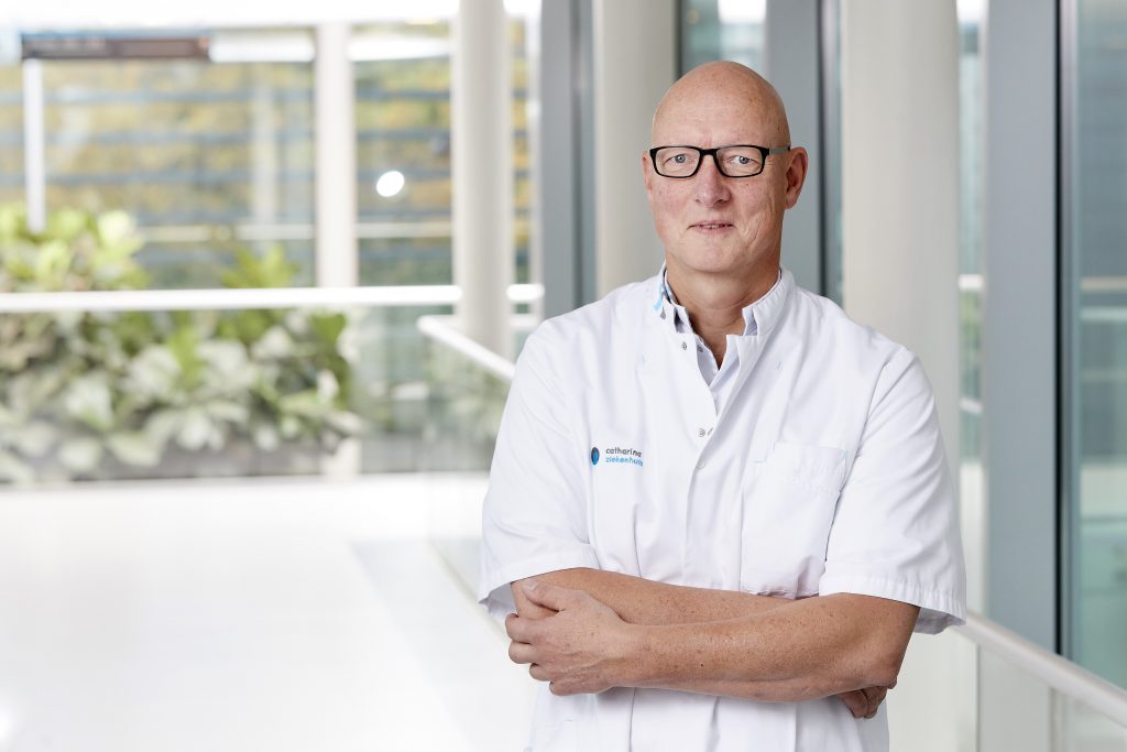 Persoon met bril en witte artsenjas staat met gekruiste armen in een lichte ruimte met grote ramen en planten op de achtergrond.