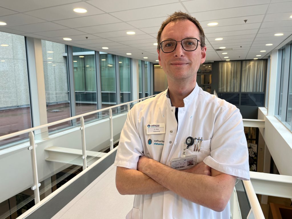 Een persoon in een witte artsenjas met naamkaartje staat met gekruiste armen in een moderne hal en kijkt naar de camera.