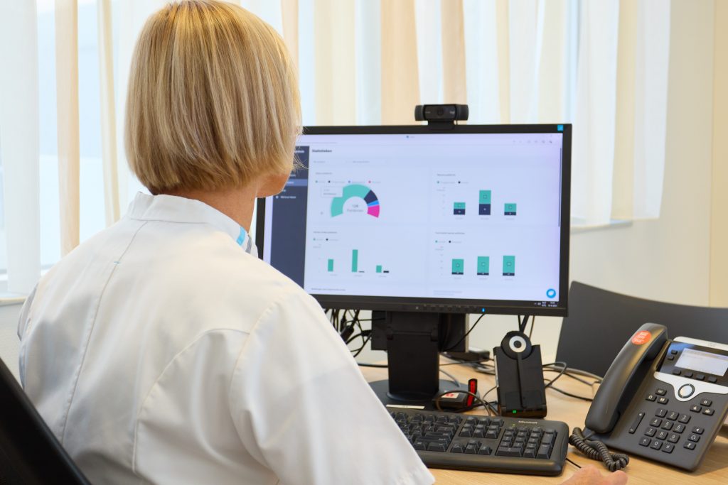 Persoon in witte labjas achter bureau kijkt naar computerscherm met COPD-thuismonitoring, telefoon op tafel.