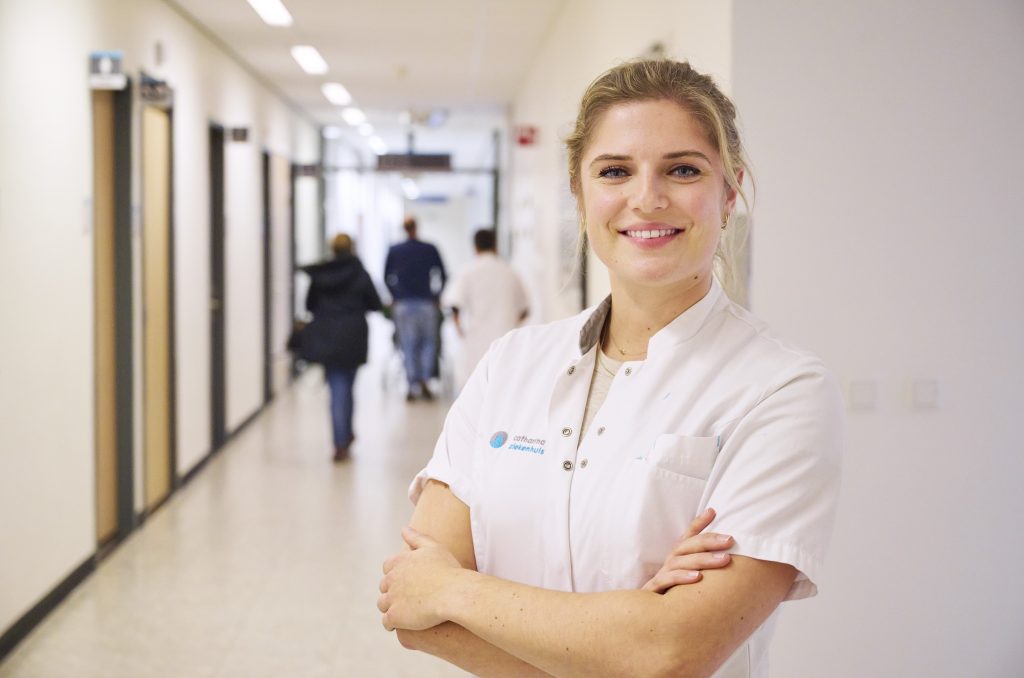 Vrouw in witte doktersjas staat met gekruiste armen en glimlacht in heldere zorggang, mensen lopen op achtergrond.